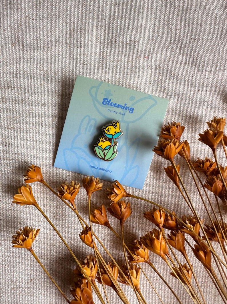 Peut inclure: Une carte bleu clair avec le texte "Blooming Bunny Bud" et une &eacute;pingle lapin. L'&eacute;pingle repr&eacute;sente un lapin jaune avec un masque bleu. Des fleurs orange s&eacute;ch&eacute;es sont dispos&eacute;es autour de la carte.