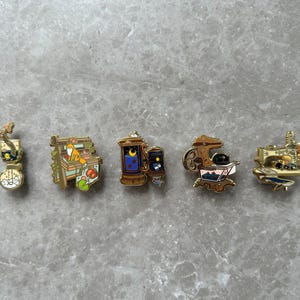 Puede incluir: Una colección de cinco pines de esmalte con varios diseños. Los pines incluyen un bonsái, un gato en una estantería, una linterna con una luna y estrellas, una cámara de cine con un gato y una máquina de coser. Los pines son dorados y multicolores.