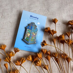 Peut inclure: &Eacute;pingle en &eacute;mail bleue repr&eacute;sentant une lanterne avec un chat &agrave; l'int&eacute;rieur, pos&eacute;e sur une carte bleu clair. La carte indique "Blooming" et "Rose Cat Enamel Pin". Des fleurs oranges s&eacute;ch&eacute;es sont dispers&eacute;es.