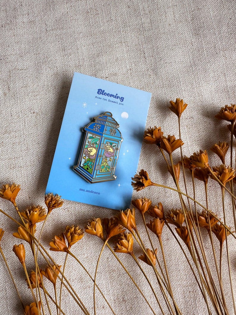 Peut inclure: &Eacute;pingle en &eacute;mail bleue repr&eacute;sentant une lanterne avec un chat &agrave; l'int&eacute;rieur, pos&eacute;e sur une carte bleu clair. La carte indique "Blooming" et "Rose Cat Enamel Pin". Des fleurs oranges s&eacute;ch&eacute;es sont dispers&eacute;es.