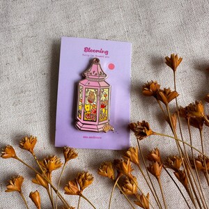 Peut inclure: &Eacute;pingle en &eacute;mail rose en forme de lanterne, avec le mot "Blooming" sur la carte de support. L'&eacute;pingle pr&eacute;sente un design d&eacute;taill&eacute; avec des motifs floraux et animaliers. Des fleurs s&eacute;ch&eacute;es entourent l'&eacute;pingle.
