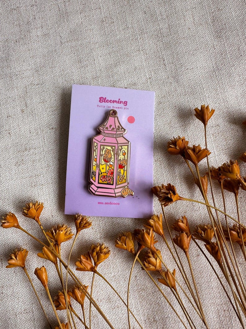 Peut inclure: &Eacute;pingle en &eacute;mail rose en forme de lanterne, avec le mot "Blooming" sur la carte de support. L'&eacute;pingle pr&eacute;sente un design d&eacute;taill&eacute; avec des motifs floraux et animaliers. Des fleurs s&eacute;ch&eacute;es entourent l'&eacute;pingle.