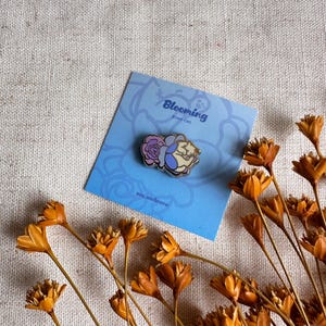 Peut inclure: &Eacute;pingle en &eacute;mail Blooming Rose Cat repr&eacute;sentant un chat avec une rose. L'&eacute;pingle est pos&eacute;e sur une carte bleue avec le texte "Blooming Rose Cat" et "mn.andcoco". Des fleurs orange s&eacute;ch&eacute;es sont dispos&eacute;es autour de la carte.