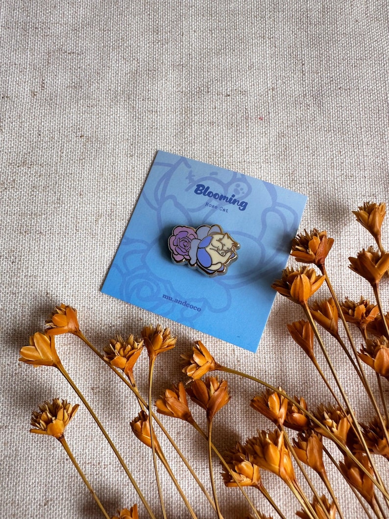 Peut inclure: &Eacute;pingle en &eacute;mail Blooming Rose Cat repr&eacute;sentant un chat avec une rose. L'&eacute;pingle est pos&eacute;e sur une carte bleue avec le texte "Blooming Rose Cat" et "mn.andcoco". Des fleurs orange s&eacute;ch&eacute;es sont dispos&eacute;es autour de la carte.
