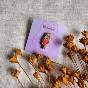 Peut inclure: &Eacute;pinglette Blooming Dolly Cat sur une carte lavande. L'&eacute;pinglette repr&eacute;sente un chat avec des accents rouges et oranges. La carte porte le texte "Blooming Dolly Cat". Des fleurs orange s&eacute;ch&eacute;es sont dispos&eacute;es autour de la carte.