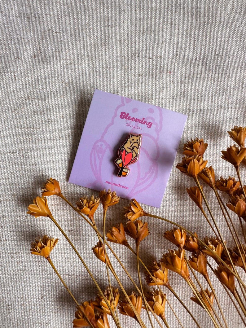 Peut inclure: &Eacute;pinglette Blooming Dolly Cat sur une carte lavande. L'&eacute;pinglette repr&eacute;sente un chat avec des accents rouges et oranges. La carte porte le texte "Blooming Dolly Cat". Des fleurs orange s&eacute;ch&eacute;es sont dispos&eacute;es autour de la carte.