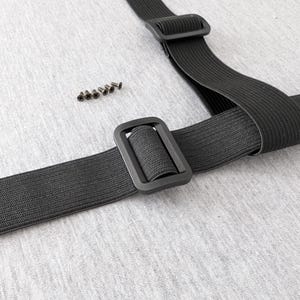 Pu&ograve; includere: Cinturino elastico nero con fibbia rettangolare e diverse piccole viti. Il cinturino &egrave; ripiegato e presenta una superficie strutturata. L'hardware &egrave; anch'esso nero e sembra essere realizzato in plastica o materiale simile. Lo sfondo &egrave; un tessuto grigio chiaro.