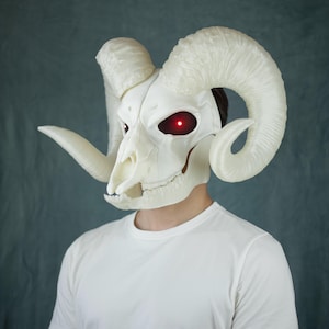 Op de afbeelding: Een wit ramsschedelmasker met grote, gekrulde hoorns en oplichtende rode LED-ogen. Het masker bedekt het bovenste deel van het gezicht en het hoofd, met gedetailleerde botachtige texturen en een realistische skeletstructuur.