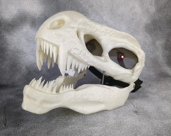 May include: A white plastic dinosaur skull mask with a detailed texture and sharp teeth. The mask has a black strap for wearing.