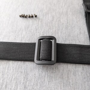 Pu&ograve; includere: Primo piano di un cinturino elastico nero con fibbia rettangolare. Un piccolo mucchio di rivetti metallici &egrave; sopra il cinturino. Il cinturino e la fibbia sono su uno sfondo grigio chiaro.