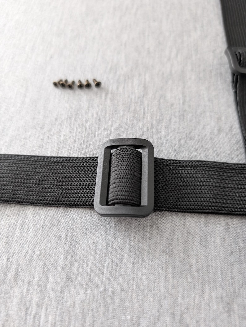 Pu&ograve; includere: Primo piano di un cinturino elastico nero con fibbia rettangolare. Un piccolo mucchio di rivetti metallici &egrave; sopra il cinturino. Il cinturino e la fibbia sono su uno sfondo grigio chiaro.