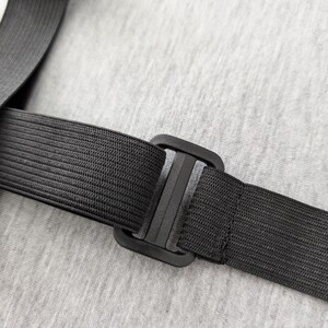 Pu&ograve; includere: Primo piano di un cinturino elastico nero con fibbia in plastica. Il cinturino &egrave; strutturato e sembra far parte di un oggetto pi&ugrave; grande. Lo sfondo &egrave; un tessuto grigio chiaro.