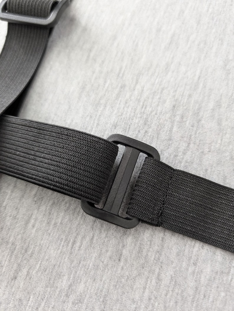 Pu&ograve; includere: Primo piano di un cinturino elastico nero con fibbia in plastica. Il cinturino &egrave; strutturato e sembra far parte di un oggetto pi&ugrave; grande. Lo sfondo &egrave; un tessuto grigio chiaro.
