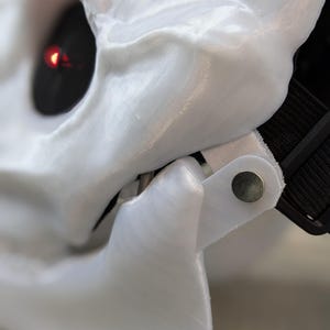 May include: Close-up of a white plastic skull mask with a red light in the eye socket. The mask has a black strap and a silver metal hinge.