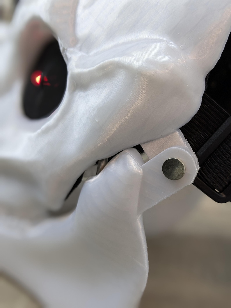May include: Close-up of a white plastic skull mask with a red light in the eye socket. The mask has a black strap and a silver metal hinge.