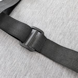 Pu&ograve; includere: Primo piano di un cinturino elastico nero con una fibbia di plastica. Il cinturino &egrave; realizzato in materiale tessuto ed &egrave; collegato a un altro cinturino. Lo sfondo &egrave; un tessuto grigio chiaro.