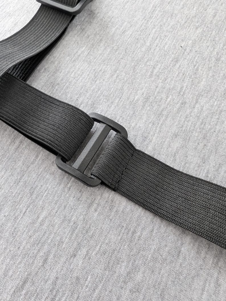 Pu&ograve; includere: Primo piano di un cinturino elastico nero con una fibbia di plastica. Il cinturino &egrave; realizzato in materiale tessuto ed &egrave; collegato a un altro cinturino. Lo sfondo &egrave; un tessuto grigio chiaro.