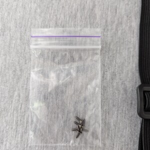 Pu&ograve; includere: Un piccolo sacchetto di plastica trasparente con chiusura a zip viola contiene diverse piccole viti color argento. La busta &egrave; su una superficie di tessuto grigio strutturato. Sul lato destro dell'immagine &egrave; visibile una cinghia nera con fibbia.