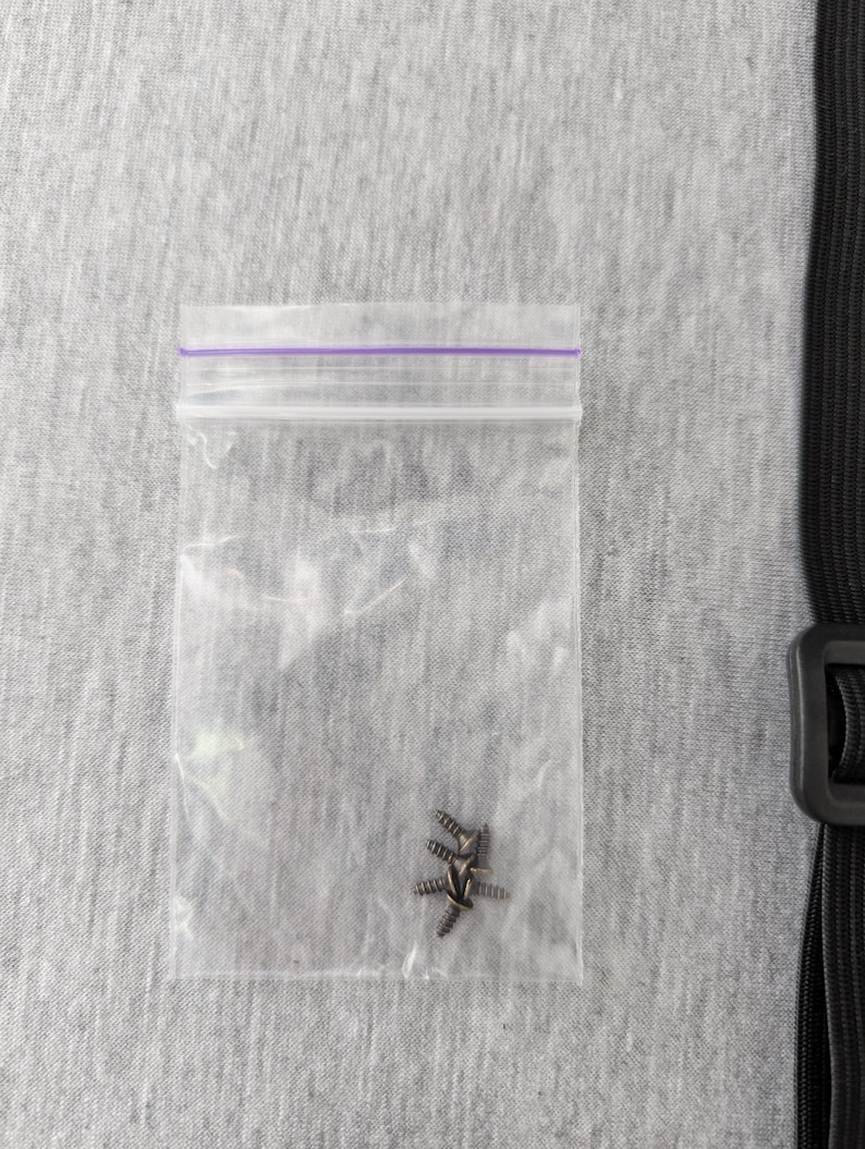 Pu&ograve; includere: Un piccolo sacchetto di plastica trasparente con chiusura a zip viola contiene diverse piccole viti color argento. La busta &egrave; su una superficie di tessuto grigio strutturato. Sul lato destro dell'immagine &egrave; visibile una cinghia nera con fibbia.