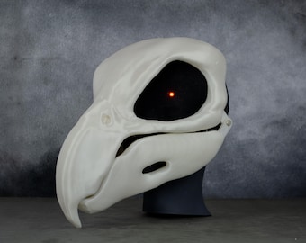 May include: A white plastic bird skull mask with a black eye socket and a red light inside. The mask is on a black stand.