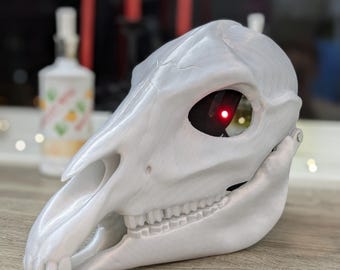 Pode incluir: Uma máscara de crânio de animal branca impressa em 3D com uma luz vermelha na órbita.