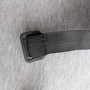 Pu&ograve; includere: Primo piano di una cintura in nylon nero con fibbia rettangolare. La cintura &egrave; realizzata in materiale tessuto ed &egrave; attaccata a una fibbia in plastica nera. Lo sfondo &egrave; un tessuto grigio chiaro.