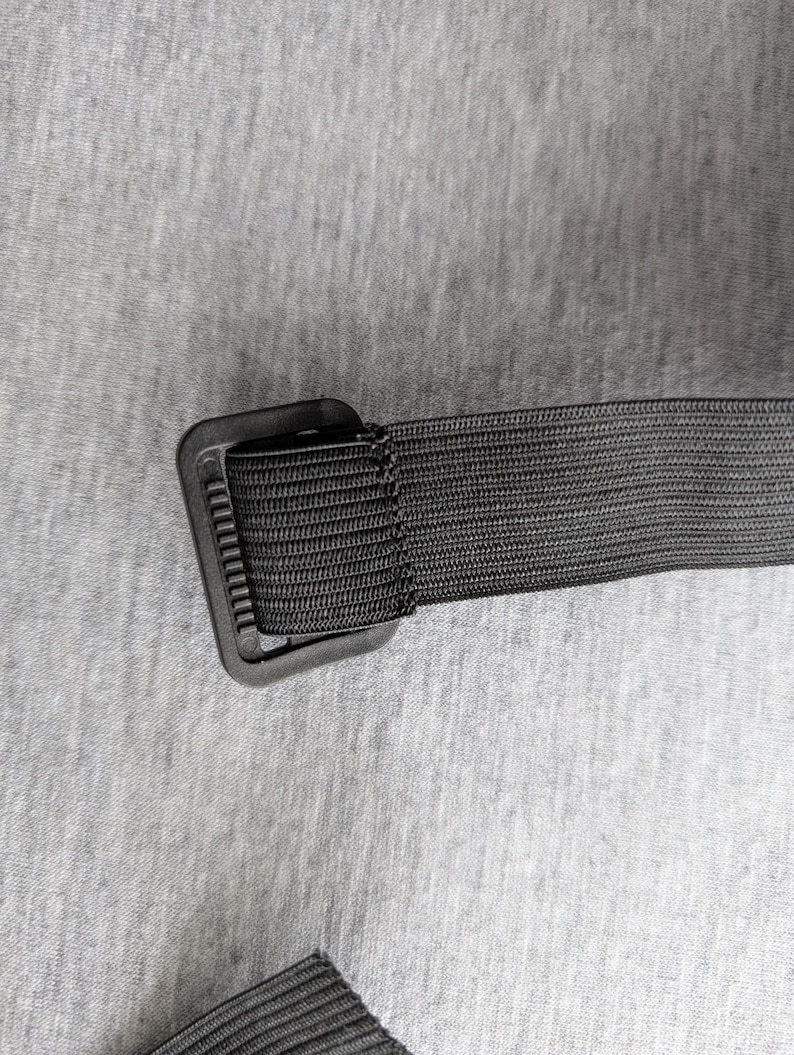 Pu&ograve; includere: Primo piano di una cintura in nylon nero con fibbia rettangolare. La cintura &egrave; realizzata in materiale tessuto ed &egrave; attaccata a una fibbia in plastica nera. Lo sfondo &egrave; un tessuto grigio chiaro.