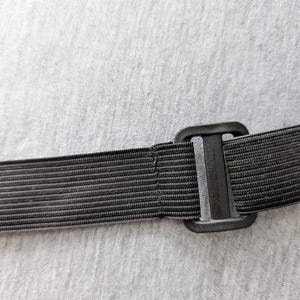Pu&ograve; includere: Primo piano di un cinturino elastico nero con una fibbia di plastica. Il cinturino &egrave; a coste e sembra far parte di un articolo pi&ugrave; grande. Lo sfondo &egrave; un tessuto grigio chiaro.