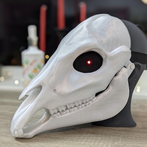 May include: A white, 3D printed animal skull mask with a red LED eye. The mask is detailed with teeth and a textured surface.