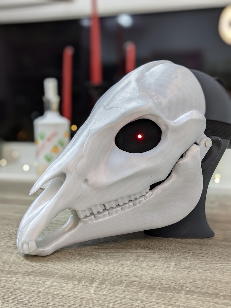 May include: A white, 3D printed animal skull mask with a red LED eye. The mask is detailed with teeth and a textured surface.