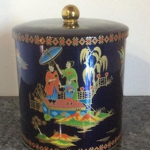 Może przedstawiać: Cylindryczne, granatowe pudełko z pokrywką i złotym guzikiem. Pudełko zdobi kolorowa scena inspirowana Azją z postaciami, parasolem i mostem. Obręcz i podstawa mają dekoracyjny złoty i pomarańczowy wzór.