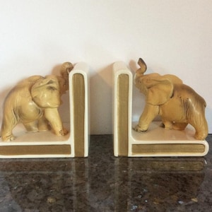 Może przedstawiać: Para podpórka do książek z figurką beżowego słonia po obu stronach. Figurki słoni stoją na białej podstawie z złotym wzorem książki.