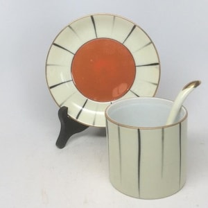 Puede incluir: Un juego de taza y platillo de cerámica blanca con rayas negras y doradas. El platillo tiene un círculo naranja grande en el centro. La taza tiene un diseño a juego y viene con una cucharita.