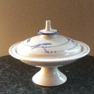 May include: A white ceramic covered dish with a pedestal base. The lid has a blue and white dragonfly design. The dish is on a dark gray surface.