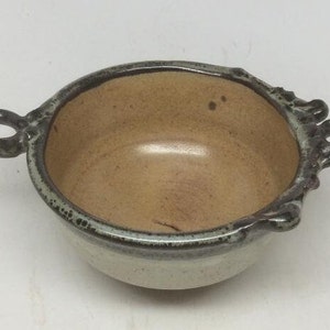 May include: A small, light brown ceramic bowl with a light green glaze and two handles. The bowl has a rustic, handmade look.