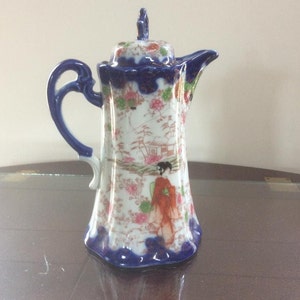 Può includere: Una teiera in porcellana bianca con un motivo floreale blu e oro. La teiera ha un manico e un coperchio. La teiera è decorata con una scena di una donna in kimono rosso.