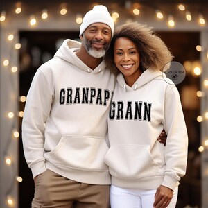 May include: A couple wearing matching cream-colored hoodies with the words "GRANPAP" and "GRANN" in black block letters. The man wears a white beanie. The woman has curly hair and is wearing white pants.