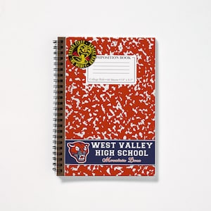 May include: A red composition notebook with a white speckled pattern. The notebook has a brown spiral binding and a sticker of a cobra with the words "Strike First Strike Hard No Mercy" on it. There is also a sticker of a mountain lion with the words "West Valley High School Mountain Lions" on it.