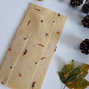 May include: Three beige reusable food wraps with a fall-themed print of carrots, beets, and leaves. The wraps are folded on a white surface with fall-themed decorations.