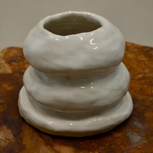 Puede incluir: Un jarrón de cerámica blanca con un diseño apilado y redondeado. El jarrón tiene una abertura ancha y se asienta sobre una superficie de madera. La cerámica tiene un acabado mate y una superficie ligeramente texturizada.