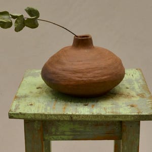 May include: A small, rustic, brown ceramic vase with a narrow neck, holding a sprig of greenery, sits on a small, weathered, green wooden stool. The stool has a square top and four legs. The vase is approximately 15 cm tall.
