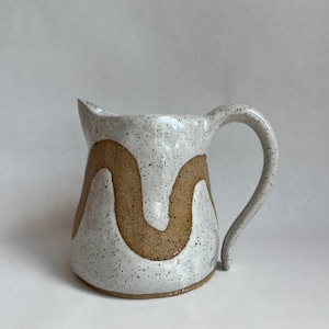 May include: A ceramic jug with a white speckled glaze and a brown wavy pattern. The jug features a curved handle and a pouring spout. The design is a modern, minimalist style, suitable for serving drinks.