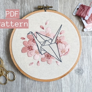 May include: A PDF pattern for an embroidery hoop featuring a pink origami crane surrounded by pink cherry blossoms. The text "PDF Pattern" is in the top left corner of the image.