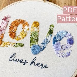 Puede incluir: Arte de aro bordado con la palabra "LOVE" en diseños florales coloridos. La palabra "love" está cosida en un arcoíris de colores, con "lives here" en azul oscuro debajo. El texto "PDF Pattern" está en un cuadro rosa.