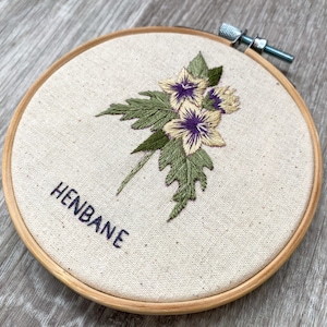 May include: Embroidered hoop art featuring a cluster of yellow and purple flowers with green leaves. The text "HENBANE" is embroidered below the flowers.