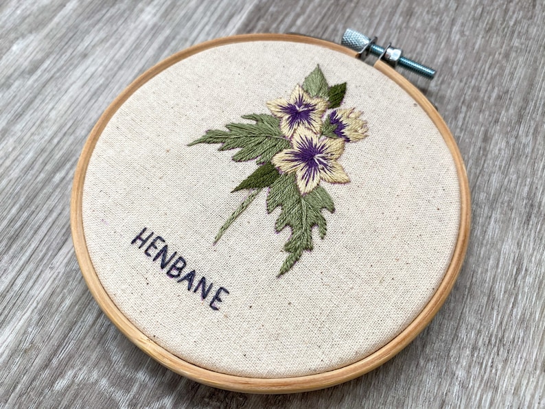 May include: Embroidered hoop art featuring a cluster of yellow and purple flowers with green leaves. The text "HENBANE" is embroidered below the flowers.