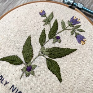 May include: Embroidered hoop art featuring a green plant with purple flowers and the text "DEADLY NIGHT" at the bottom.