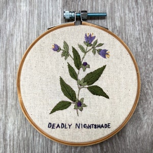 May include: Embroidered hoop art featuring a green plant with purple flowers and the text "DEADLY NIGHTSHADE".