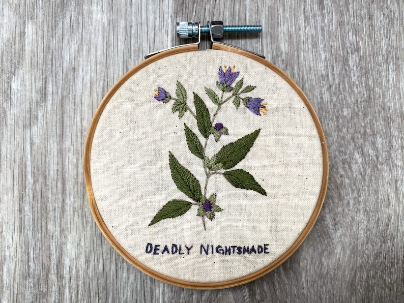 May include: Embroidered hoop art featuring a green plant with purple flowers and the text "DEADLY NIGHTSHADE".