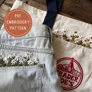 Könnte beinhalten: Hellblaue Jeans mit gestickten weißen Gänseblümchen in der Tasche. Eine Tragetasche aus Segeltuch mit Trader Joe's-Logo und passender Gänseblümchen-Stickerei befindet sich neben der Jeans. Das Bild enthält auch ein PDF-Stickmuster.
